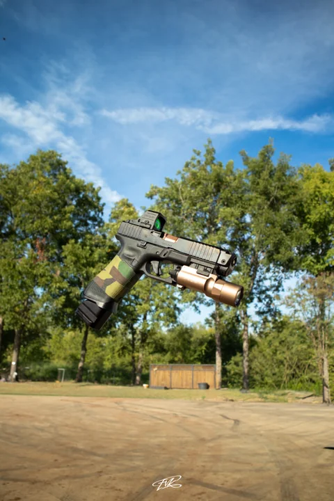Handgun with camouflage grip photographed against trees and a blue sky