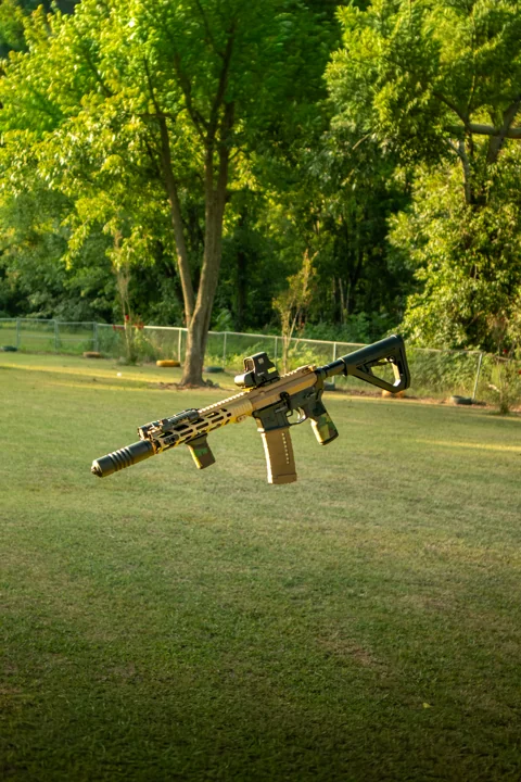 Tan and black rifle photographed against a sunlit lawn and trees